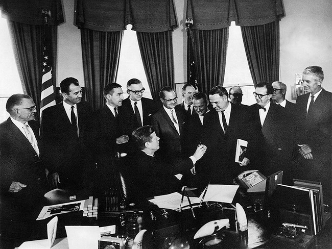President John F. Kennedy hands a pen to Congressman Clem Miller during the signing ceremony for the Point Reyes National Seahore's Enabling Legislation on September 13, 1962. In an ornate office, eleven men dressed in suits look on from behind a large desk as a man seated at the desk turns in his chair to hand a pen to a man standing to the left of him.