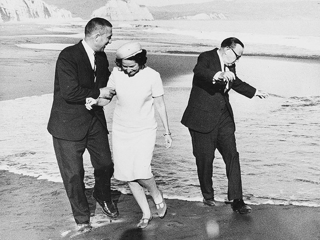 Two men wearing suits and a woman wearing a dress and a hat attempt to avoid an incoming wave at a sandy beach.