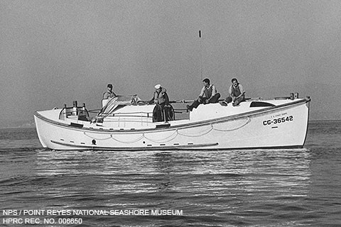 A black and white photo of four lifeboat station crew members aboard a 36-foot-long white lifeboat on gentle water.