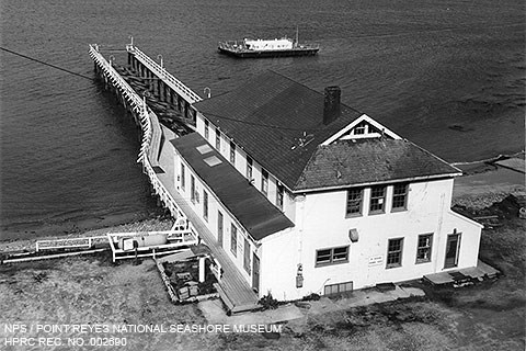 A black and white photograph of a two-story, white-sided building from which a dock extends into a bay. A small barge is anchored near the end of the dock.