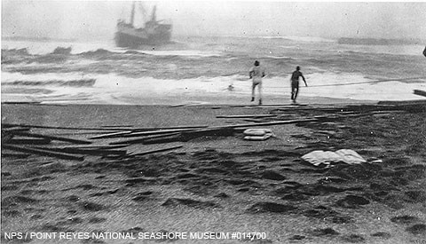 Black and white photo of two men on a beach with a ship caught in heavy surf very close to shore.
