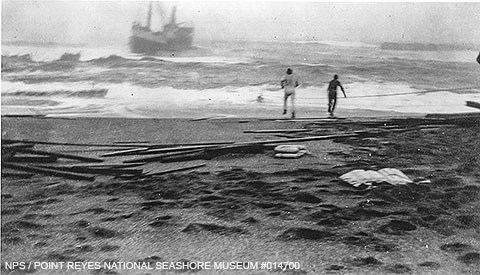Black and white photo of two men on a beach with a ship caught in heavy surf very close to shore.