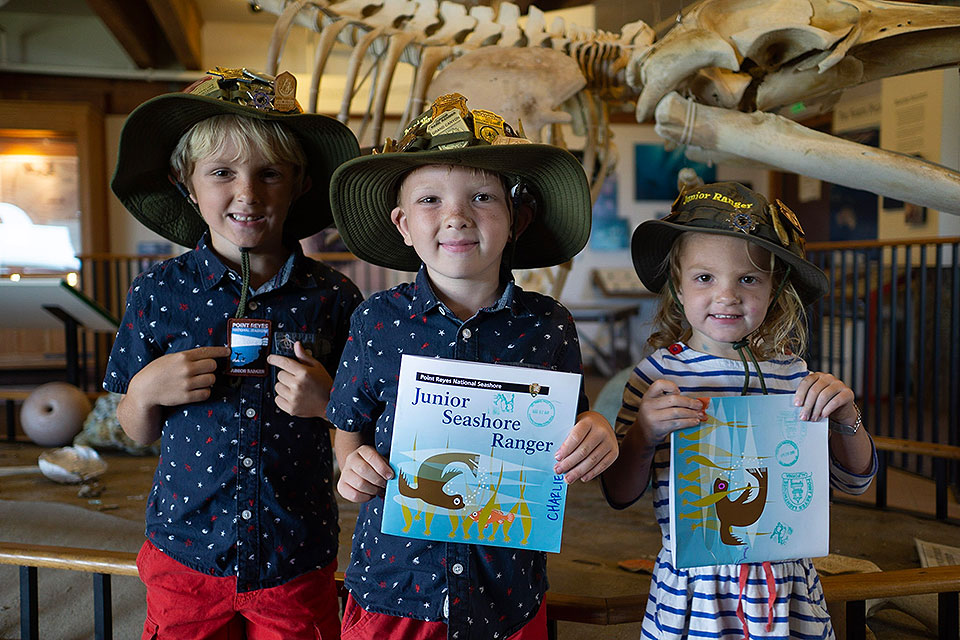 Two young boys and a young girl wearing junior ranger hats pose while holding junior ranger badges and booklets.