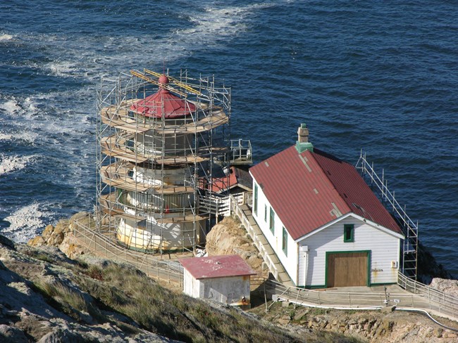 Three white-sided, red-roofed buildings perched on a rocky headland with the ocean in the background. The lighthouse tower is surrounded by scaffolding.