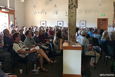 Dozens of people sitting on chairs in a large white-walled room.