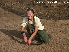 A young woman with braided blonde hair and wearing a yellow shirt kneels down to plant a seedling.