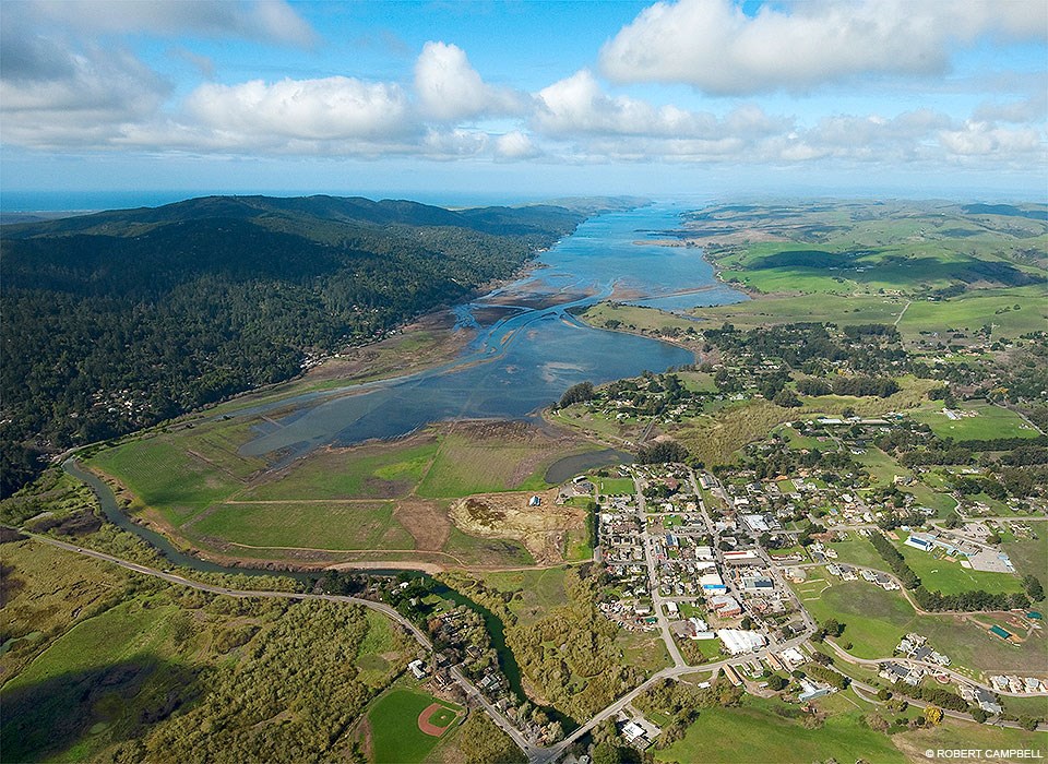 Aerial photo of recently restored wetlands. A narrow bay stretches off in the distance. A wooded ridge borders the wetlands and bay on the left. A small town is located in the lower right.