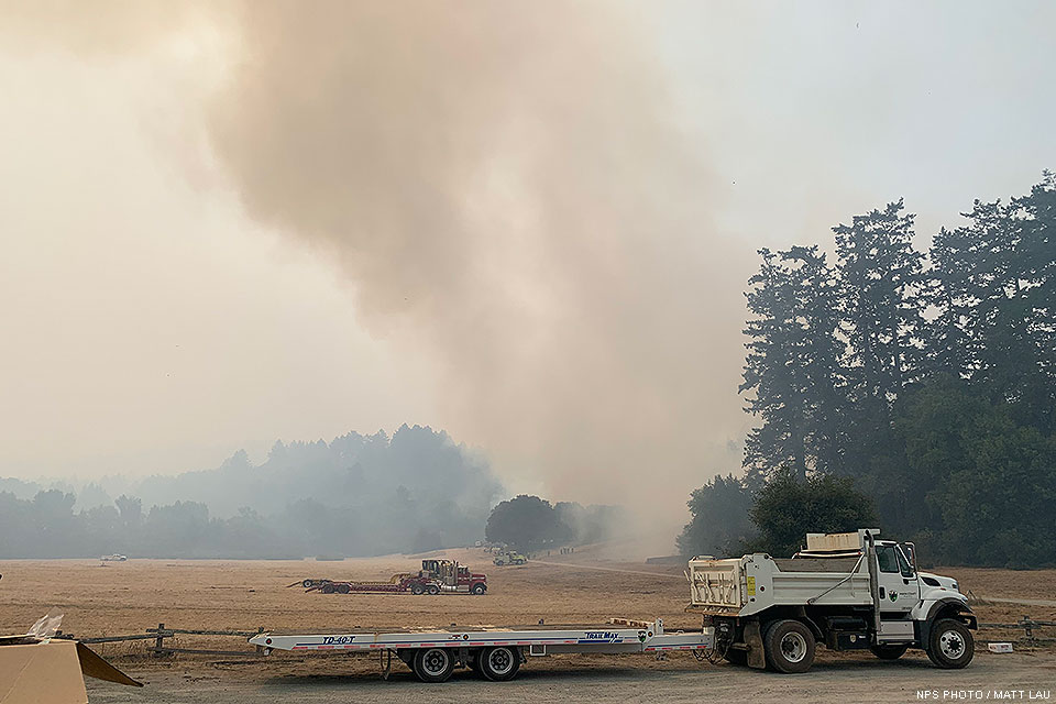 Gray smoke rises from a controlled burn along a meadow in the distance beyond some tall trees located on the right. A dump truck pulling a flatbed trailer sits in the foreground.