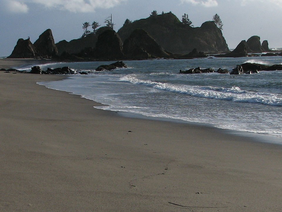 Waves wash up on a sandy ocean beach. Rocky headlands rise above the far end of the beach.