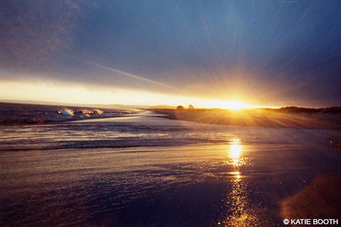 The setting sun casts rays from a gap between low clouds and the horizon, illuminating wet sand and waves washing across a sandy beach.