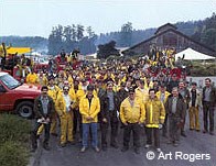 A photo of dozens of wildland firefighters wearing mostly yellow clothing standing in a parking lot with a barn-like visitor center and a forested ridge in the background.