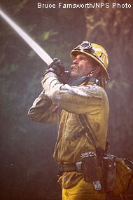 A wildland firefighter wearing yellow clothing and hat aiming a stream of water from a hose up and to the left.