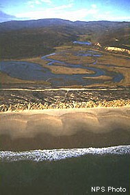 A low-altitude aerial photo of a sandy spit, an esturary, and hills shortly after a wildfire burned the area's vegetation.