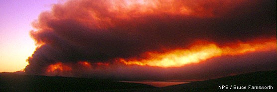 Smoke rises from a ridge on the left and drifts to the right as light from the sunrise in the distance filters through the smoke.