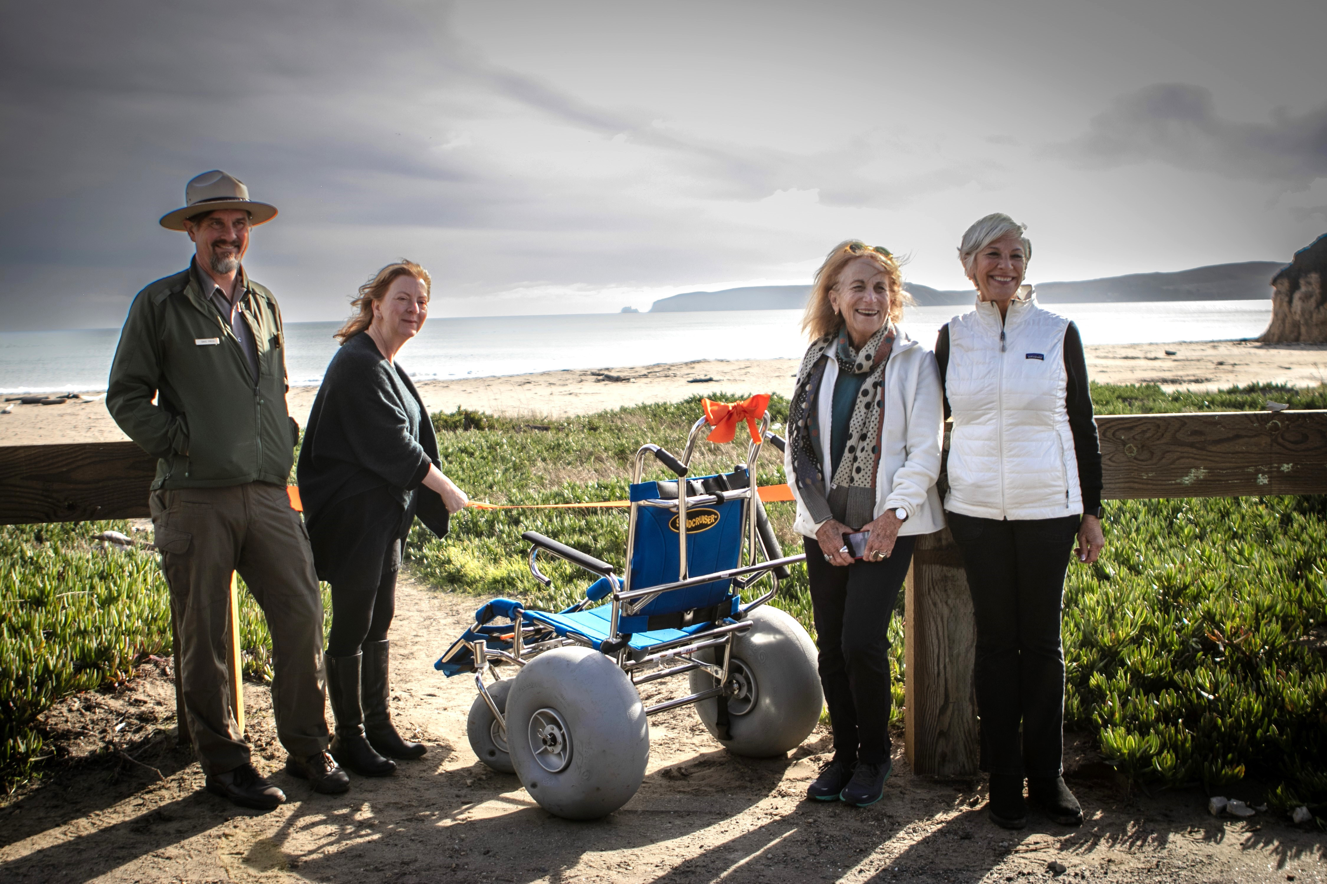 The Beach Wheelchair Ribbon Cutting Ceremony at Drakes Beach on November 9, 2023. A park ranger with three women stand beside a beach wheelchair with a beach and a bay in the background.