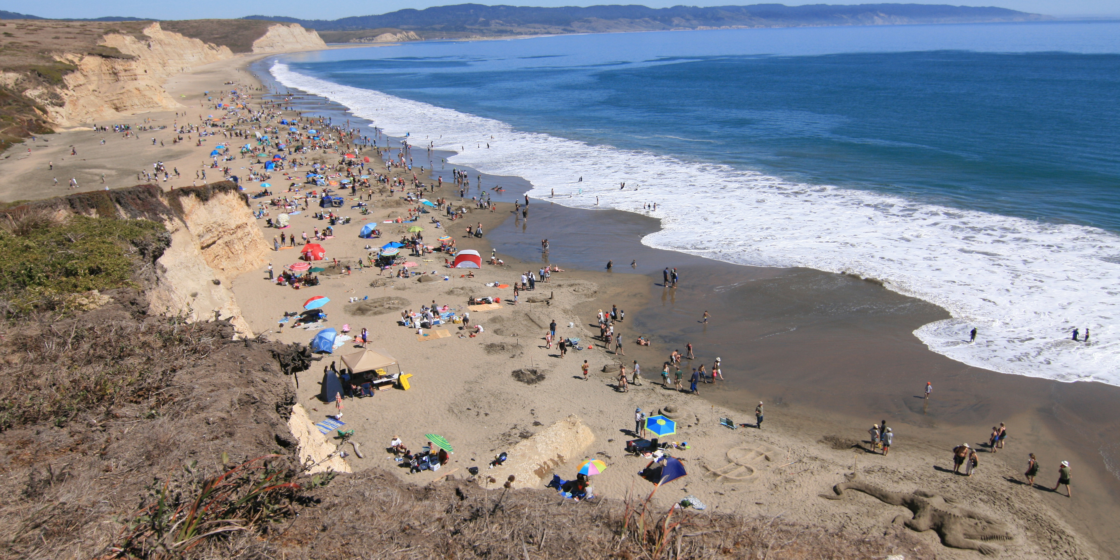 Drakes Beach during a Sand Sculpture Contest. A photo from a bluff top looking down at a crowded beach on the left with a blue ocean bay on the right.