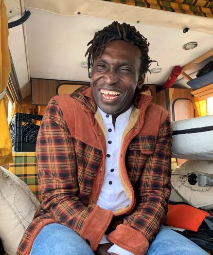 Felix Louis N'jai. A black man wearing a plaid jacket laughs while sitting in a small camper van.