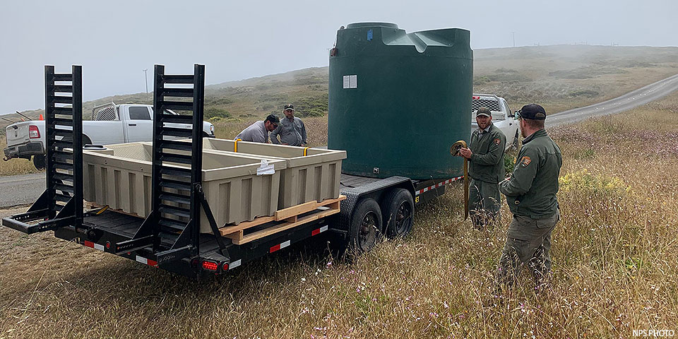 Four men in gray and green uniforms prepare to unload a large green water tank and two beige water troughs from a flatbed trailer.