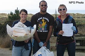 Three Beach Cleanup Volunteers holding litter collected from a beach.