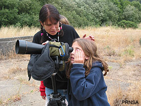 A woman assists a young girl who looks through a spotting scope.