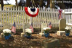 Four small white headstones in a small cemetery surrounded by a white picket fence on which some bunting is hung.