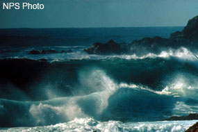 Large wave breaking on a beach with a rock outcrop, the ocean, and blue sky in the background.