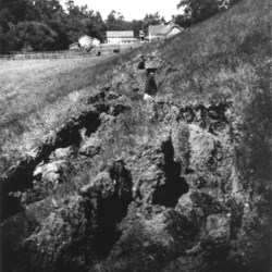 Black and white photo of a woman standing in a surface rupture, where the soil has been turned over, with a barn in the background.