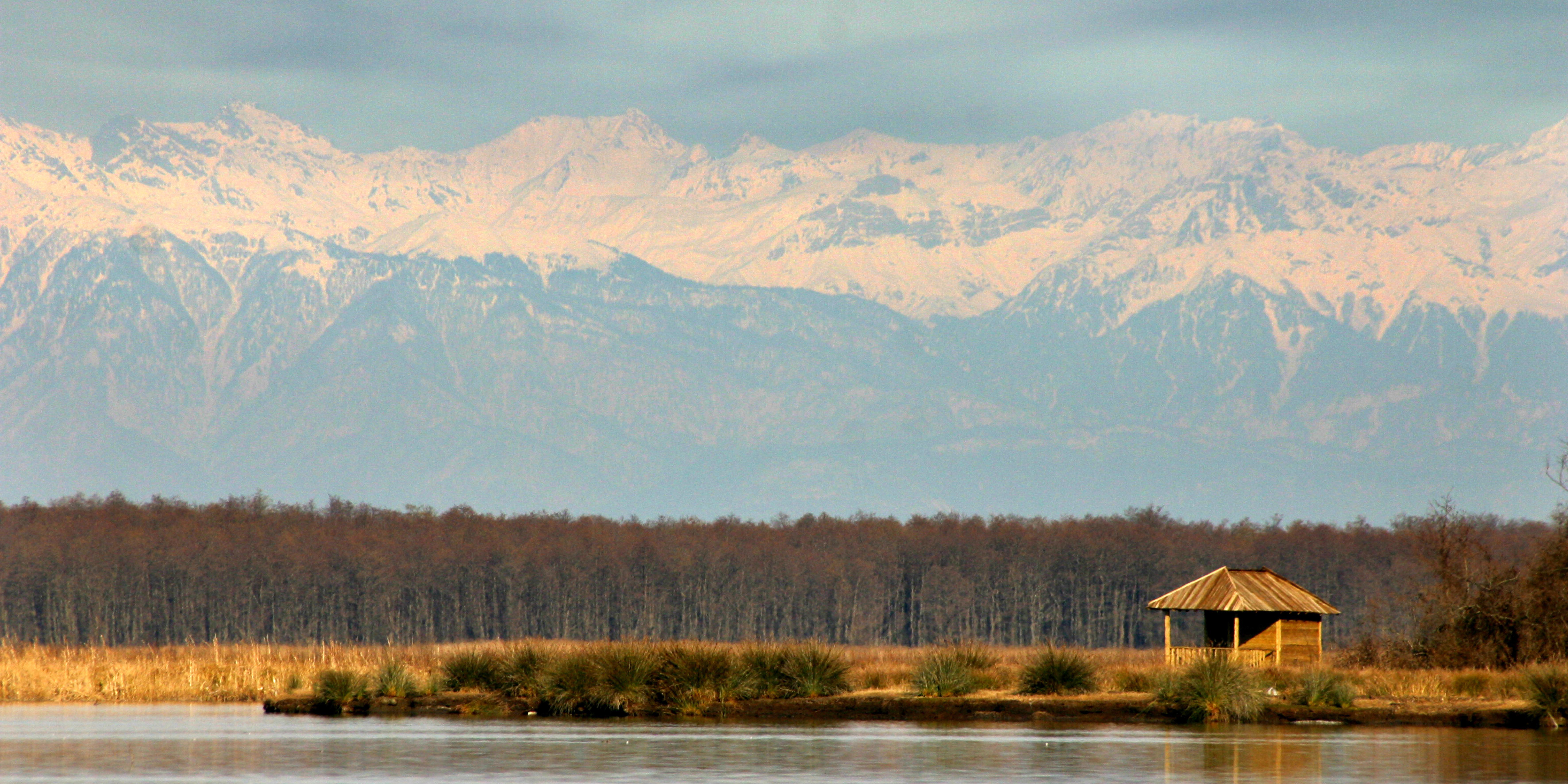 A small wooden shelter sits on a lake shore with woods and snow-capped mountains in the background.