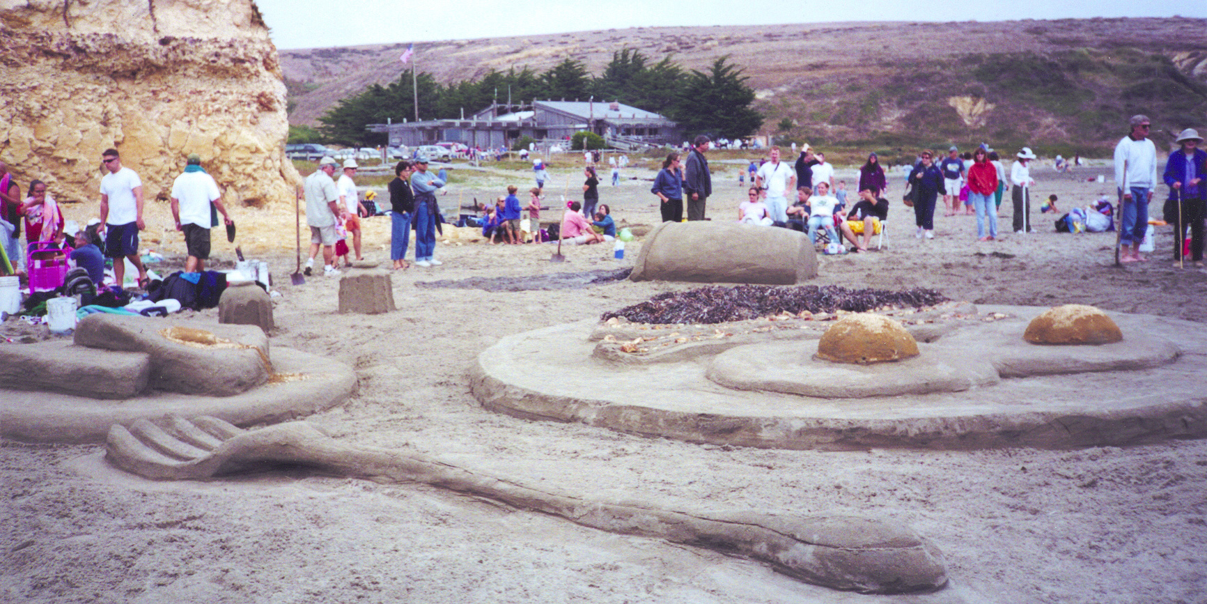 A large sand sculpture featuring a plate with two sunny side up eggs and bacon surrounded by plate with two slices of toast with butter, a fork, salt and pepper shakers, and a spilled cup of coffee