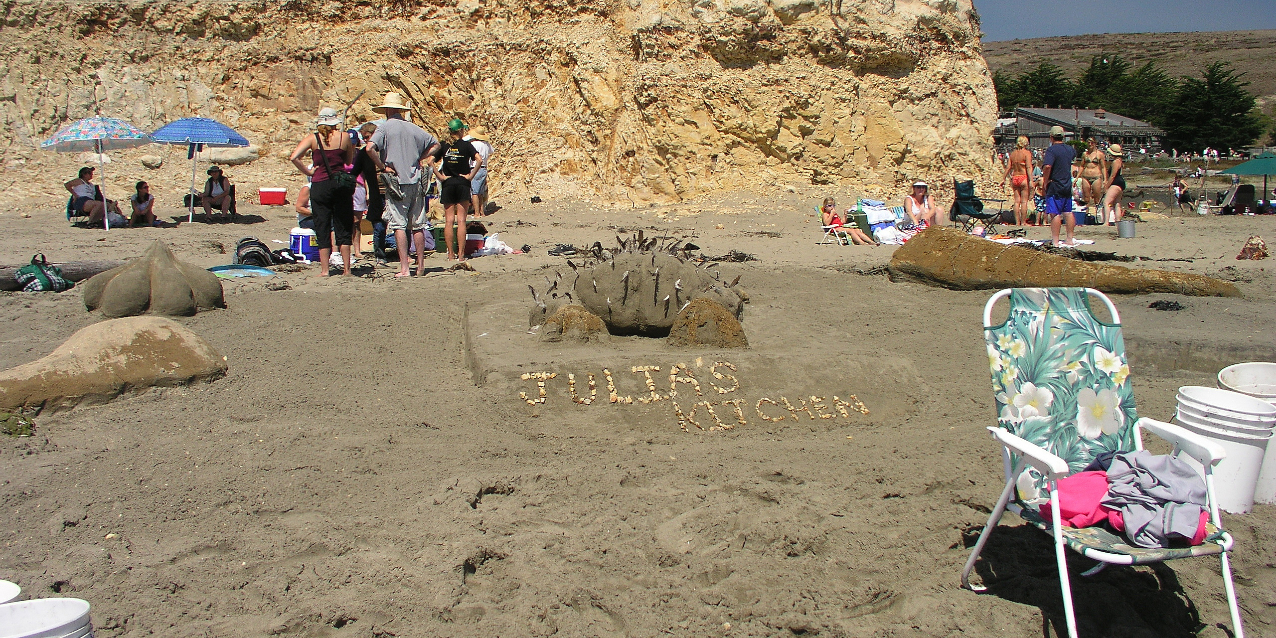 A large sand sculpture featuring a partially-plucked chicken on a flat surface labeled "Julia's Kitchen" adjacent to chicken drumsticks and a dumpling.