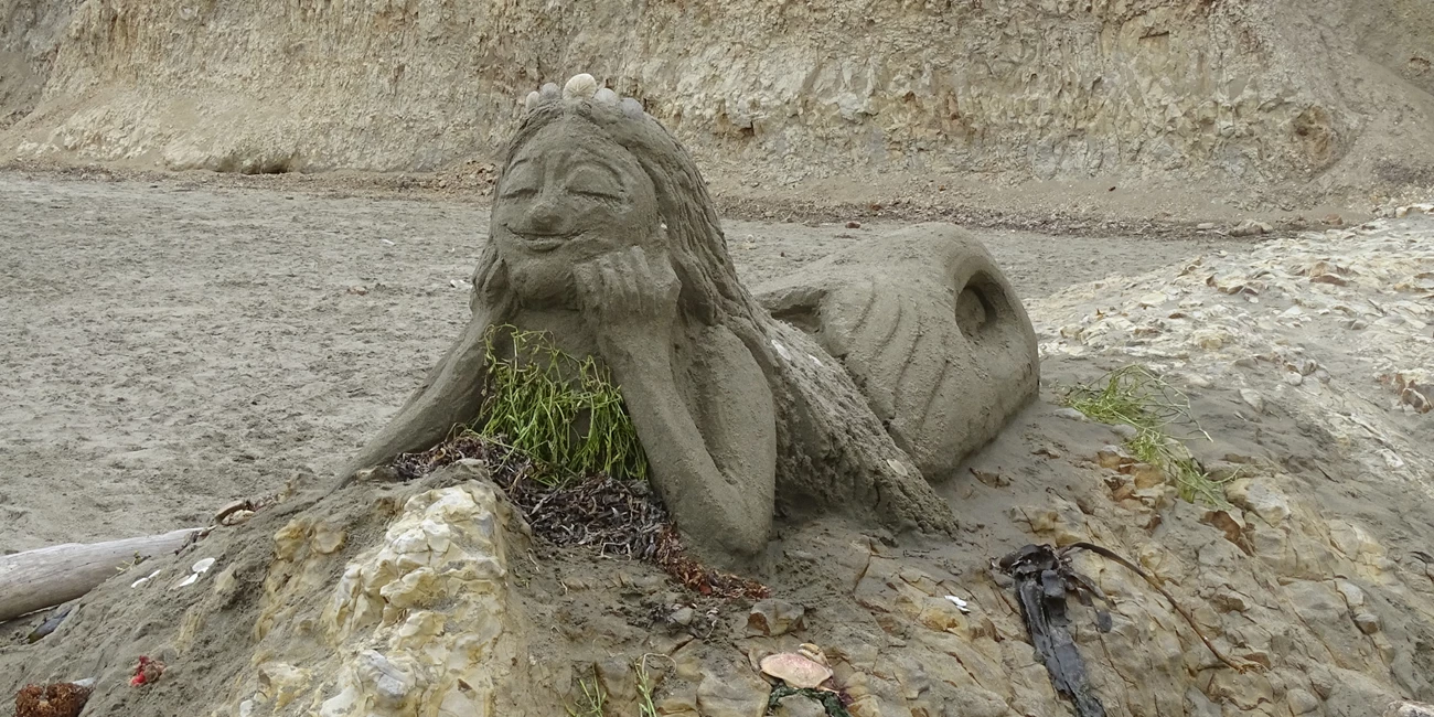 2025 Sand Sculpture Contest: Adult Category Entry #3: Maggie the Mermaid!, by Samantha and Nicole Garriott and Len and Sandy. August 23, 2025. A sand sculpture of a mermaid lying on her belly on top of a rock outcrop with her elbows on the rock and head propped up in her hands.