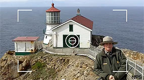 A ranger stands in the lower right foreground with lighthouse buildings and the ocean in the background.