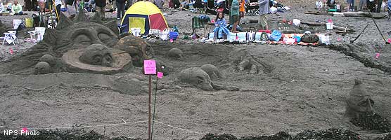 A sand sculpture of an anthropomorphic sun with its arms embracing eight anthropomorphic planets with a dwavish-looking planet outside the sun's arms.