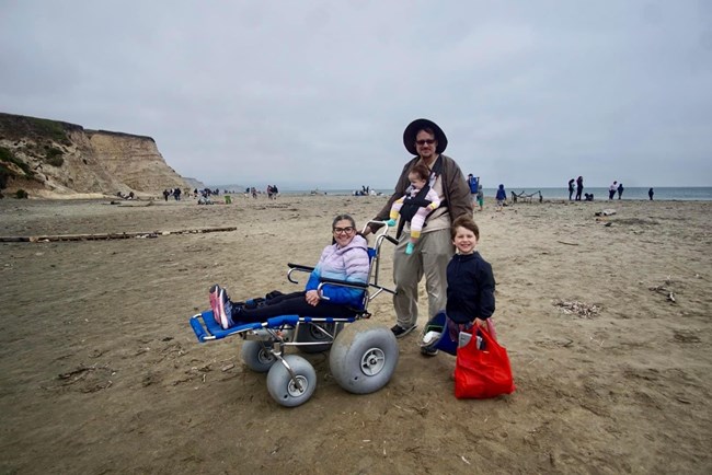 A woman in a beach wheelchair in front of a man holding a baby and a young boy, on a cloudy beach day.