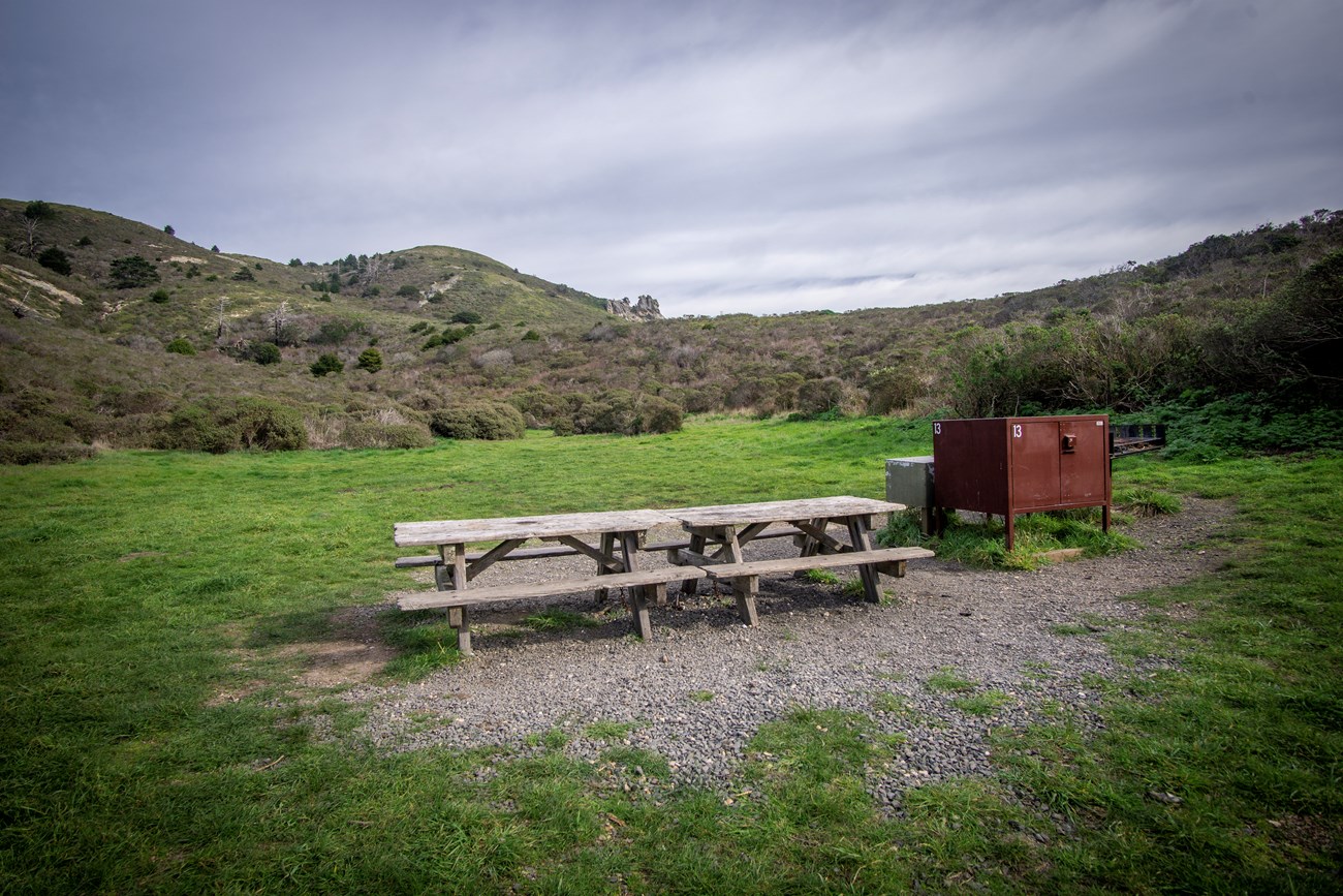 A picnic table and food storage locker in a grassy campsite amongst shrub covered hills.