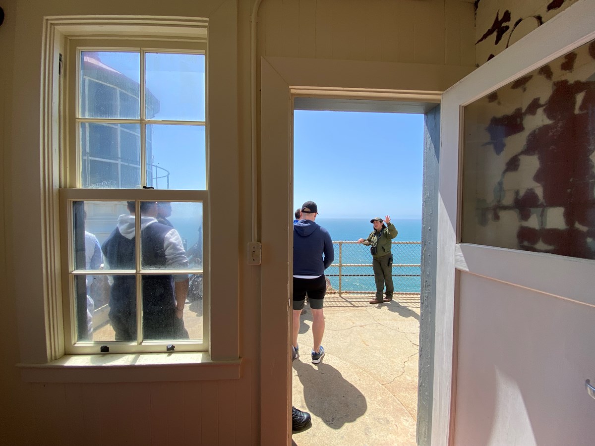 Framed by a doorway, a small group of people watches a park ranger gesture in front of the open ocean.