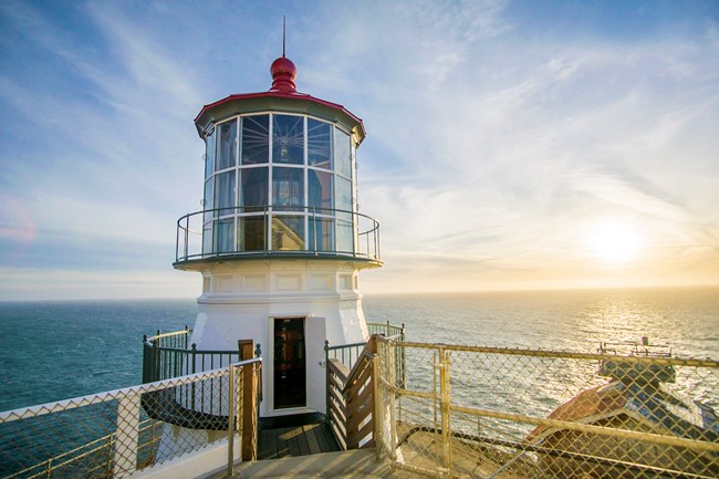 A door into a short white lighthouse with a red roof on an oceanside cliff.