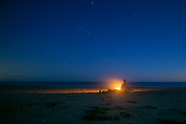 A night-time photo of a man sitting next to a small wood fire on a beach under starry skies.