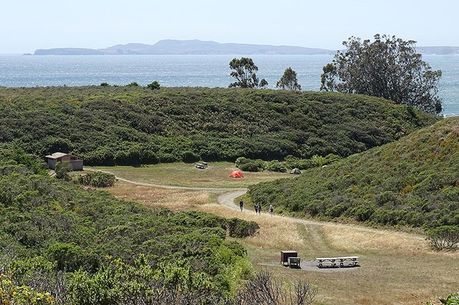 Looking down at a few campsites in a small grassy seaside valley surrounded by shrub-covered hills.