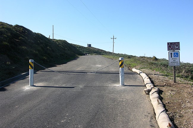 Two metal bollards connected by a heavy chain block a narrow road leading up a hill.