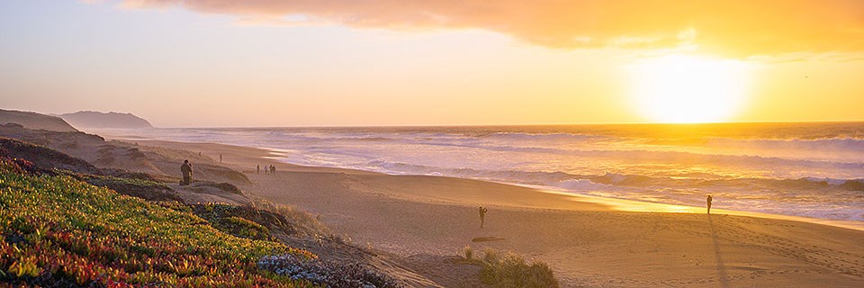 The sun sets over large waves breaking on a long, straight, sandy beach. A rocky headland rises on the left in the background.