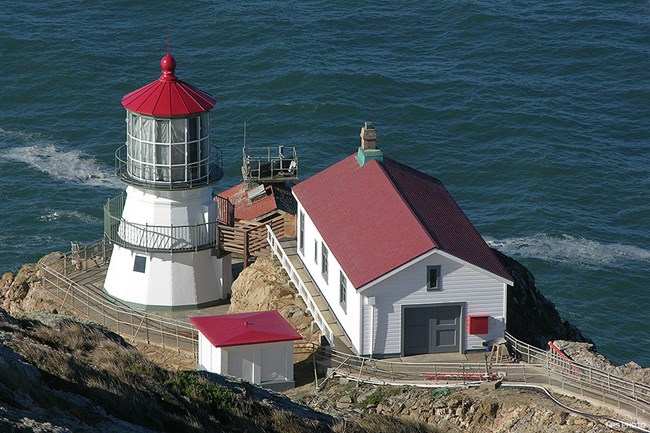 Four white-sided, red-roofed light station buildings sit on the edge of an ocean-side bluff.