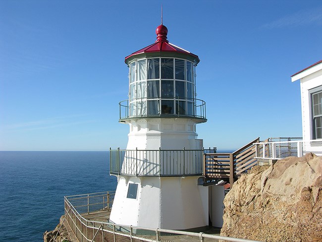 A three-story-tall, white-sided, red-roofed lighthouse perched on a rocky headland with the ocean in the background.