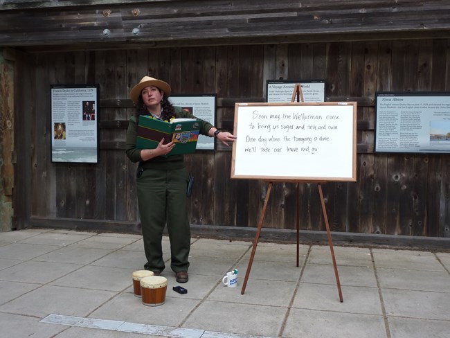 A female ranger holding a green binder and pointing to lyrics on a white board set on an easel.