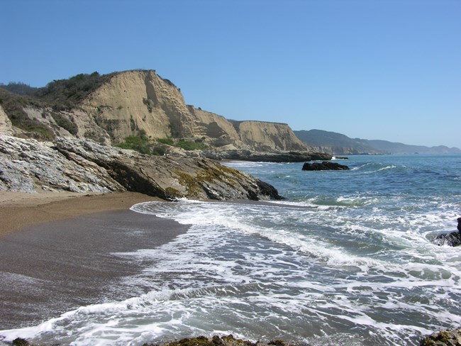 Rocky outcrops extend from tan-colored bluffs on the left into the ocean on the right.