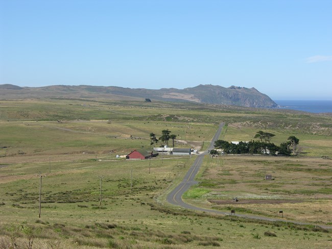 A narrow road winds through tan and green pastureland, through a cluster of buildings, and off toward a rocky headland on the edge of the ocean.