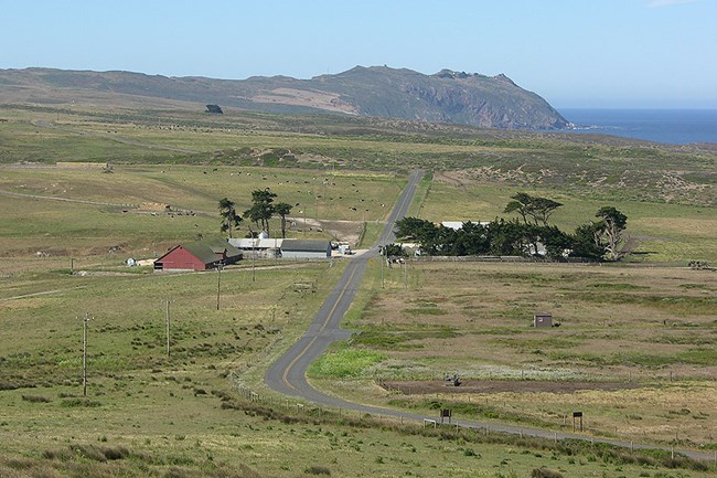 A narrow road winds through tan and green pastureland, through a cluster of buildings, and off toward a rocky headland on the edge of the ocean.