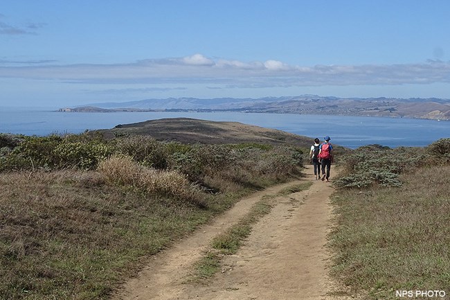 Two hikers walk along a wide dirt trail that follows a ridge separating two bodies of water that merge in the distance.