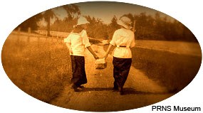 A 1920s era sepia-toned photograph of two women walking along a path through a meadow while carrying a picnic basket between them.