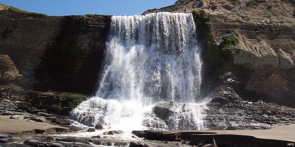 Water falling over a 30-foot tall cliff top onto a sandy beach.